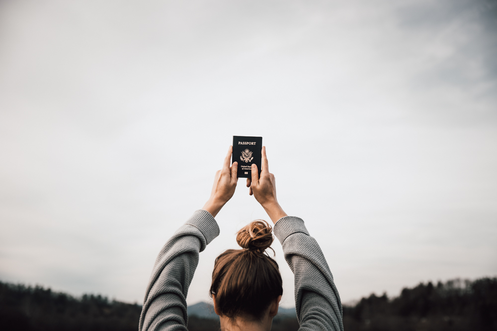 Girl holds a passport up over her head. Her hear is in a topknot and there are mountains in the background.