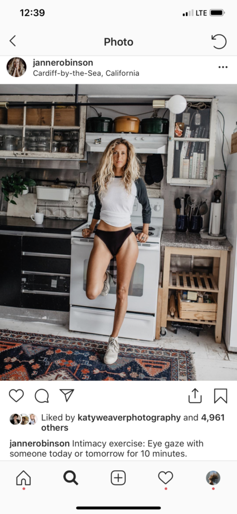 Screenshot of Janne Robinson on Instagram leaning against the stove in a kitchen in her swimsuit bottoms and long sleeve shirt, looking directly into the camera with her blonde curly hair fallen over her shoulders and half her face.